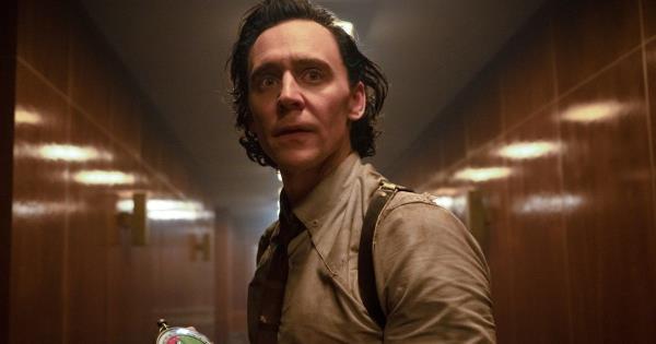 Tom Hiddleston as Loki holding a device in his hand, looking down a long hallway while wearing his tan collared shirt and suspenders in Loki.