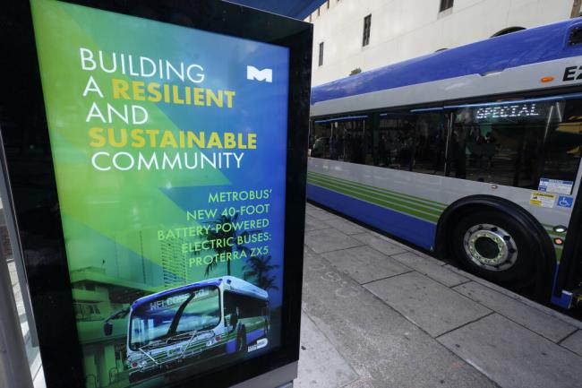 FILE - A billboard announces the benefits of Proterra ZX5 battery-powered electric buses as one is parked at a bus stop, Feb. 2, 2023, in downtown Miami. Electric bus and truck maker Proterra says it is seeking Chapter 11 bankruptcy protection from a federal court in Delaware. A statement on Proterra's website Monday, Aug 7, said that it intends to maintain normal operations and will file motions with the court to use existing capital to keep funding operations, including paying employees, vendors and suppliers. (AP Photo/Wilfredo Lee, File)